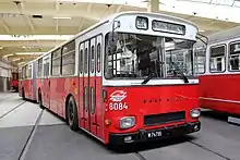 A red and white bus sits inside a brightly lit depot. It has the lightning bolt logo of the Wiener Linien, the number 8084, and the line title "26A Stadlau-Stadiobrücke"