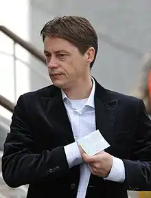 The upper body of a middle-aged white man with brown hair. He is wearing a light-coloured shirt with light blue pinstripes over a white T-shirt with a dark jacket on top. His right hand is inside his jacket and his left hand is holding a piece of paper.