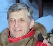 A man with grey hair, wearing a red and brown coat, with snow in the background.