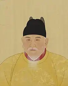 An old aged bearded man wears yellow robes with dragons inscribed and a black hat.