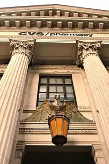 The building's entrance as seen from street level on Eighth Avenue. There is a metal lantern just above the door in the center. The door is flanked by triple-height marble columns that support a triangular pediment. The words "CVS Pharmacy" are mounted below the pediment.