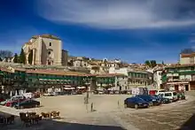 An uneven open space surrounded by houses. A church overviews the area. Cars are parked on the square.