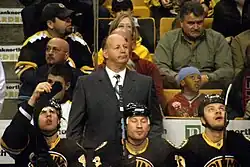 Three ice hockey players in full equipment sit at the bench during a game. Their coach is behind them watching the action on the ice.