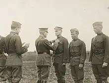 Three men in uniform are standing side by side. The one on the left is wearing a peaked "crush cap" and standing smartly at attention, while the two on the right wear garrison caps and are slouching. A man in a peaked cap and Sam Browne belt is pinning something on the chest of the first man. Behind him stands another man in a garrison cap who is reading a document in his hands.