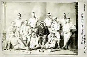 Twelve members of a baseball team are posing for a photograph, consisting of three rows; five men standing, four sitting in a chair or bench, and three sitting on the floor