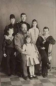 Black-and-white photograph of the Romanov family. Olga is a young girl who stands at the front resting against the arms of her seated bearded and bald father, who wears a military uniform. The older children and the empress complete the group.