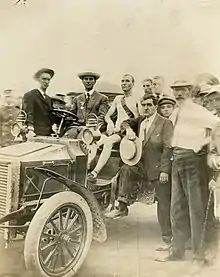 A gaunt man sits in an automobile looking exhausted, with a group of men surrounding him, looking considerably healthier. All are looking at the camera.