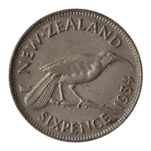 A silver coin with a huia bird perched atop a small branch. To the upper left is the label "NEW ZEALAND", to the bottom right is the label "SIXPENCE 1934"