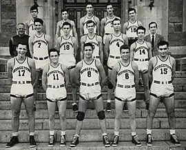 Three rows of five young men in white basketball uniforms stand on stone steps.