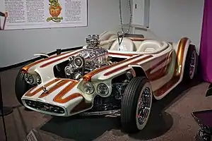 A white car with brown stripes, with open wheels and a clear bubble canopy over twin seats, and exposed, chromed engine with a blower.