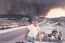 A woman looks at the camera with her mouth open, in front of a car parked on the side of the road. Behind her, thick smoke rises from a line of low hills.