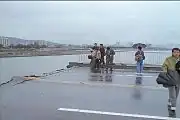 Scene from on the bridge post-collapse. Several reporters can be seen with umbrellas, and the weather is cloudy. The road is completely broken off and bent slightly upwards.