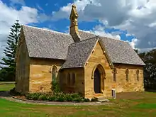 Photo of a little sandstone church with an unaisled nave, a projecting chancel, a porch, and topped with a small gabled belfry.