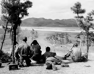 Soldiers lying in the grass with guns shooting towards a river in the distance.