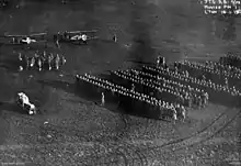 A large number of people standing in formation in a field. Two biplanes are visible at the upper left, with a smaller group of people standing in front of them.