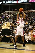 a Black athlete is shooting a basketball over another Black athlete while an Asian athlete watches.