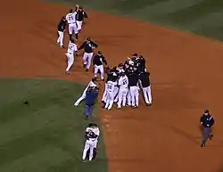 Several men in white baseball jerseys, some wearing black jackets, congregate around second base on a baseball diamond.