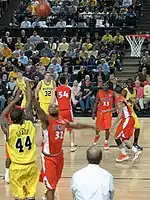 A player in a maize uniform releases a jump shot from the near corner over a defender in an orange uniform with the word Illinois on the front. Other players on both teams box out for a rebound.