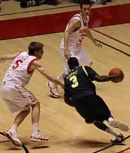 A basketball player in a dark blue uniform is dribbling past a defender in white.