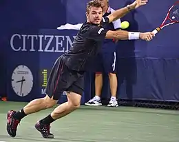 A man wearing a black shirt, black shorts, and black shoes staggers to his left to return a tennis ball as he holds his racket out on the right hand in front of his body