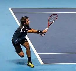 A man wearing a black shirt, black shorts, and neon yellow shoes staggers to his right to return a tennis ball as he holds his red tennis racket in front of him