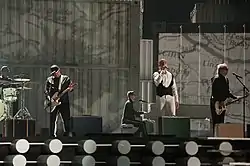 Several men in suits performing on a stage in front of a grey backdrop.