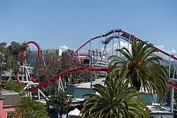 An overview image of the Flight Deck roller coaster with red track and white supports. A train is seen ascending the lift hill on the right, with the vertical loop present on the left, and the zero-gravity roll center. Various building and trees are strewn throughout the image.