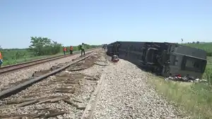 A silver train car turned on its side, with people standing on damaged railroad tracks