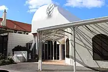 a shapley curved building single stroy entrance entrance with sunshine and shadow play on the white walls. Whimsical pink decorative columns at the entrance can be seen.