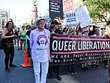 A member of the Gay Liberation Front stands beside the banner; a sign condems "rainbow merchandise" in favor of "basic civil rights," 2019