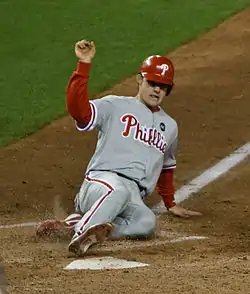 A young man in a gray baseball uniform with red trim and a red baseball helmet slides feet-first into a pentagonal white plate set in the dirt