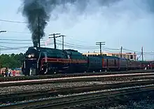 A black streamlined steam locomotive and its tender, pulling an extra tender, a Tuscan red baggage car, and a matching set of Tuscan red passenger cars