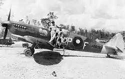 Black-and-white photo of a man wearing military uniform and two other men wearing only shorts standing on the wing of a single-engined monoplane aircraft