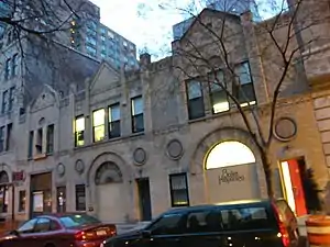 Set of 3 two-story buildings, looking mostly alike, each with a large door on the street level and a gable on the roofline, all faced in beige brick. They are adjacent to each other and to the buildings on either side. Camera is across the street from the adjacent building on the right, viewing the row in perspective.