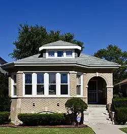 A light beige brick building with a three-sided bay to the viewer's left and a small corner porch to the viewer's right and a single dormer in the center, with a lawn that contains a bush and a small tree immediately in front of the bay.