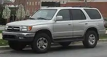 A light gray sport-utility vehicle parked on a street with brick buildings in the background, facing left