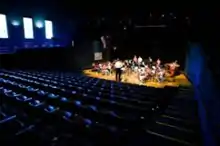  Empty blue seats and a small orchestra on a lighted stage, as seen from the rear of an auditorium