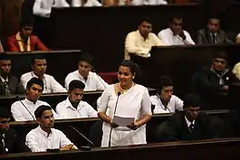 photograph of Wickramanayake dressed in a white saree surrounded by other people listening to her speech