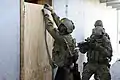 Placing an explosive breaching charge on a door, positioned to separate the door from its hinges