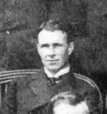 A man, fresh-faced with dark, brushed-back hair, seated among a group. He is wearing a naval officer's uniform with a high, stiff collar