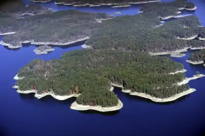 Aerial view of a calm, shiny blue lake and matriculating around wooded peninsulas covered in green, orange and red foliage