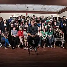 An image of a man sitting on a stool with a bunch of people behind him.