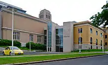 A light orange brick building with a stone and glass section connecting it to a yellow brick building, the rear of the building in the lede image. A tower rises in the distance over the structure, and there is a parking lot in front with a yellow Volkswagen New Beetle.