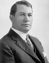 A man with dark, wavy hair, wearing a black coat, patterned tie, and white shirt