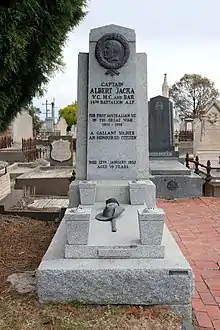 granite grave with headstone.  a brass relief showing the head of Albert Jack is at the top of the headstone.  A soldier's slouch hat and sword made of brass lay on top of the grave.