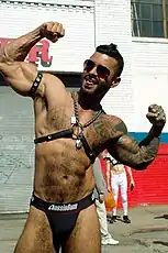 Man wearing harness and speedo, wearing sunglasses, posing and showing off his biceps, with arms raised upwards and his body slightly tilting to his left.