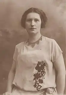 A young white woman with dark hair, wearing a light-colored short-sleeved embroidered blouse and a distinctive beaded collar necklace