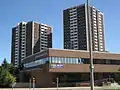 A two-storey brown brick building, with a ramp. Behind it is two high rise buildings with the same brick but additional white highlights in the centre of each side