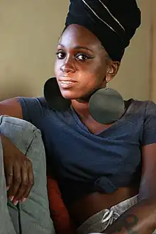 Alyesha Wise sitting on a coach, wearing a blue short-sleeve shirt rolled up at the belly, grey sweatpants and a black headwrap