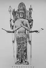 Front view of a standing statue with robes four pairs of arms. Each pair is almost left-right symmetric. Two hands are placed as in prayer with palms facing each other in front of the body. Other pairs of arms are in various positions holding flowers, a short object shaped like a flute, a necklace and long pole weapons. There is a large halo behind the head of the statue.